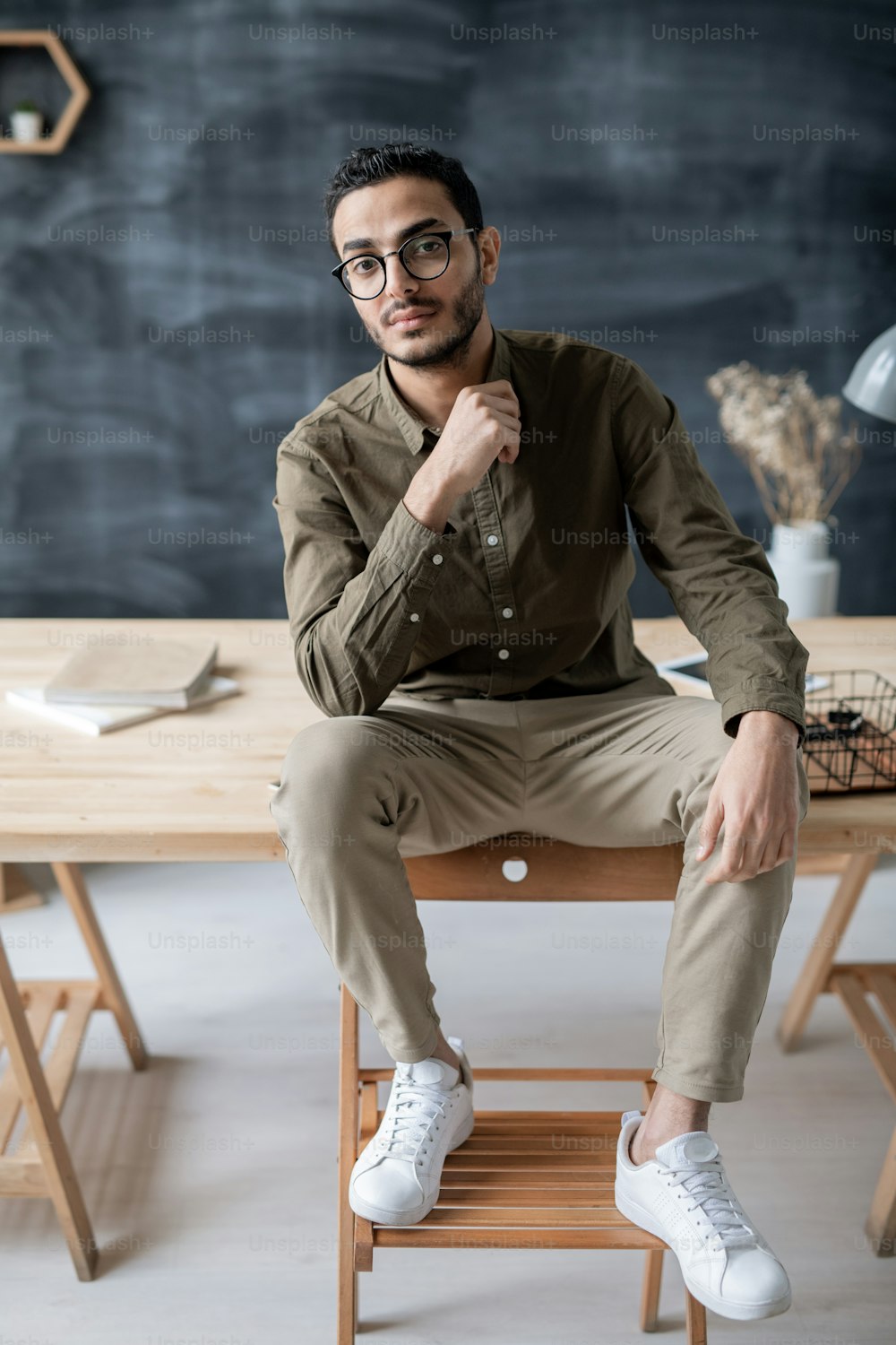 Young serious businessman sitting on wooden table with his feet on chair in front of camera and looking at you