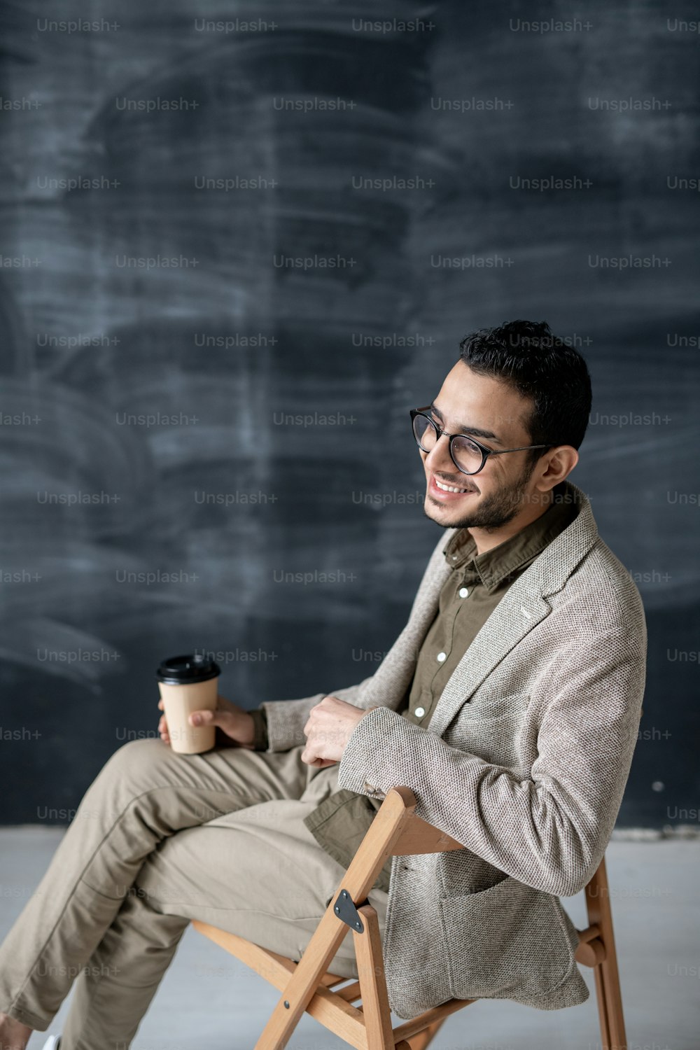 眼鏡をかけた若い陽気な男性とスマートカジュアルで、カメラの前の椅子に座って飲み物を飲む