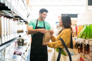 Proprietario maschio sorridente che dà sacchetto di carta al cliente femminile e vende cibo alla rinfusa al supermercato