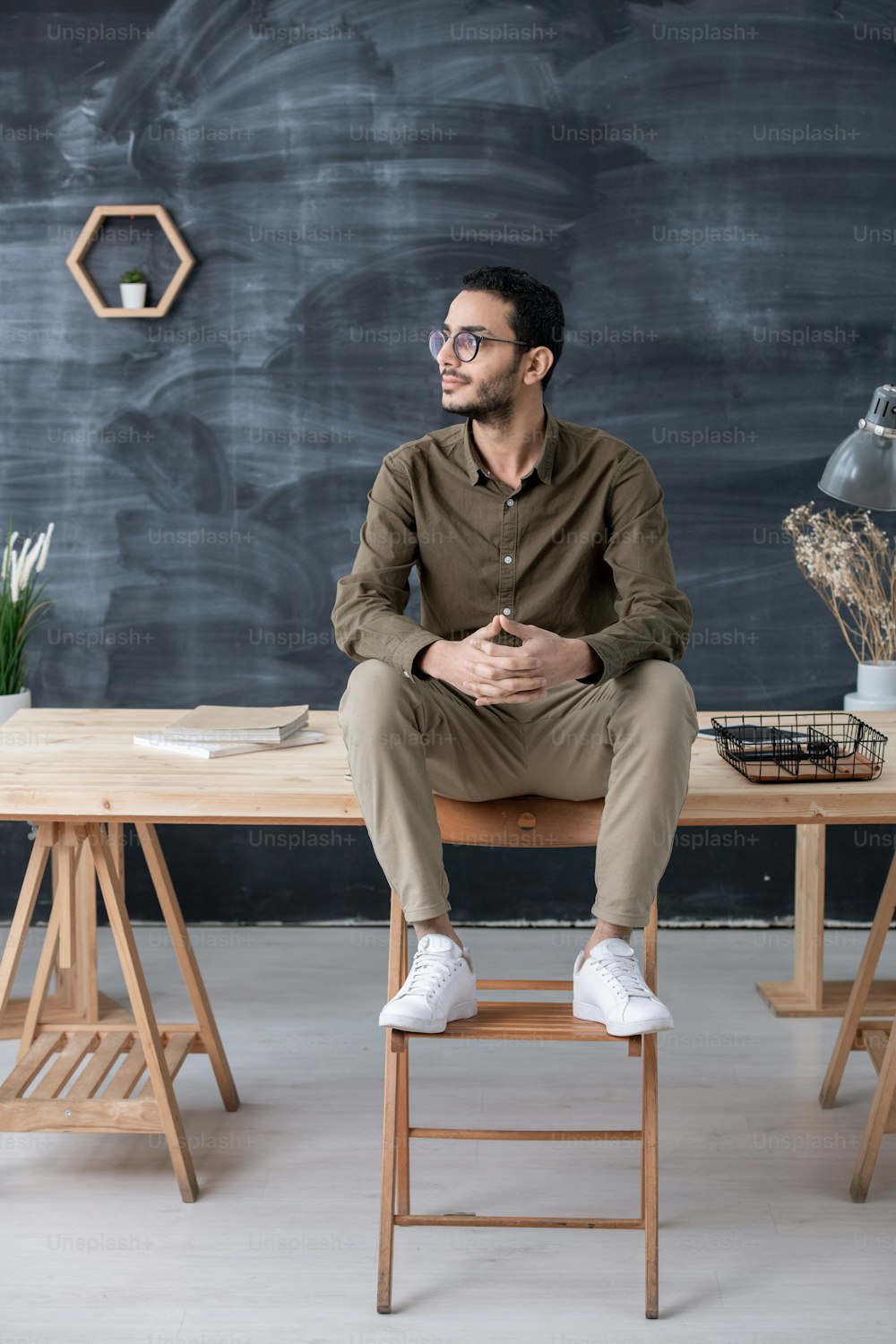 Giovane uomo d'affari riposante in abbigliamento casual seduto sul tavolo di legno con i piedi sulla sedia davanti alla telecamera