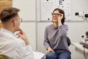 Portrait of excited young woman putting on new glasses and smiling happily in ophthalmology clinic, copy space