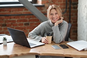 회색 스웨터를 입은 흥분한 비즈니스 레이디가 사무실의 나무 테이블에 앉아 커피를 마시고 있다