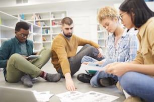 Full length view at multi-ethnic group of students sitting on floor in library and discussing project