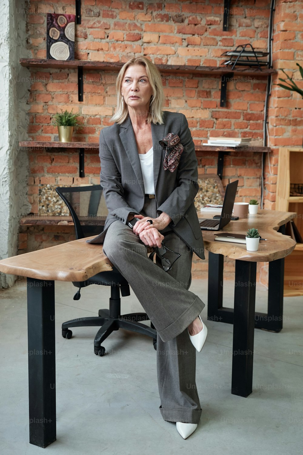 Pensativa séria mulher de negócios madura em terno elegante sentado na mesa no escritório loft