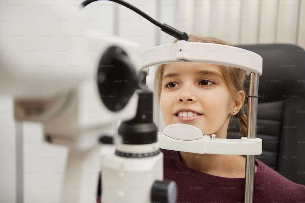 Portrait of cute smiling girl looking into refractometer during vision test in modern ophthalmology clinic, copy space