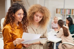 Two young businesswomen looking at documents, their colleagues coworking behind them