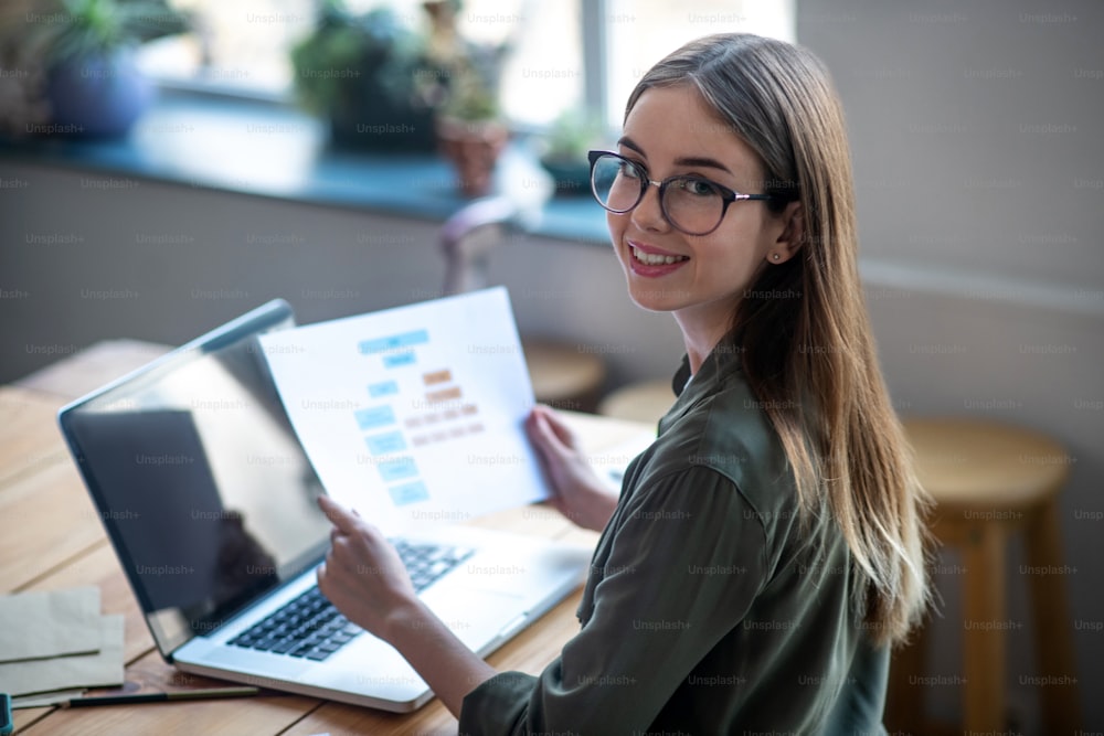 Analysis of the work done. Girl with glasses sitting at a table in front of a laptop holding a diagram in her hands, smiling and happy, looking to the side.