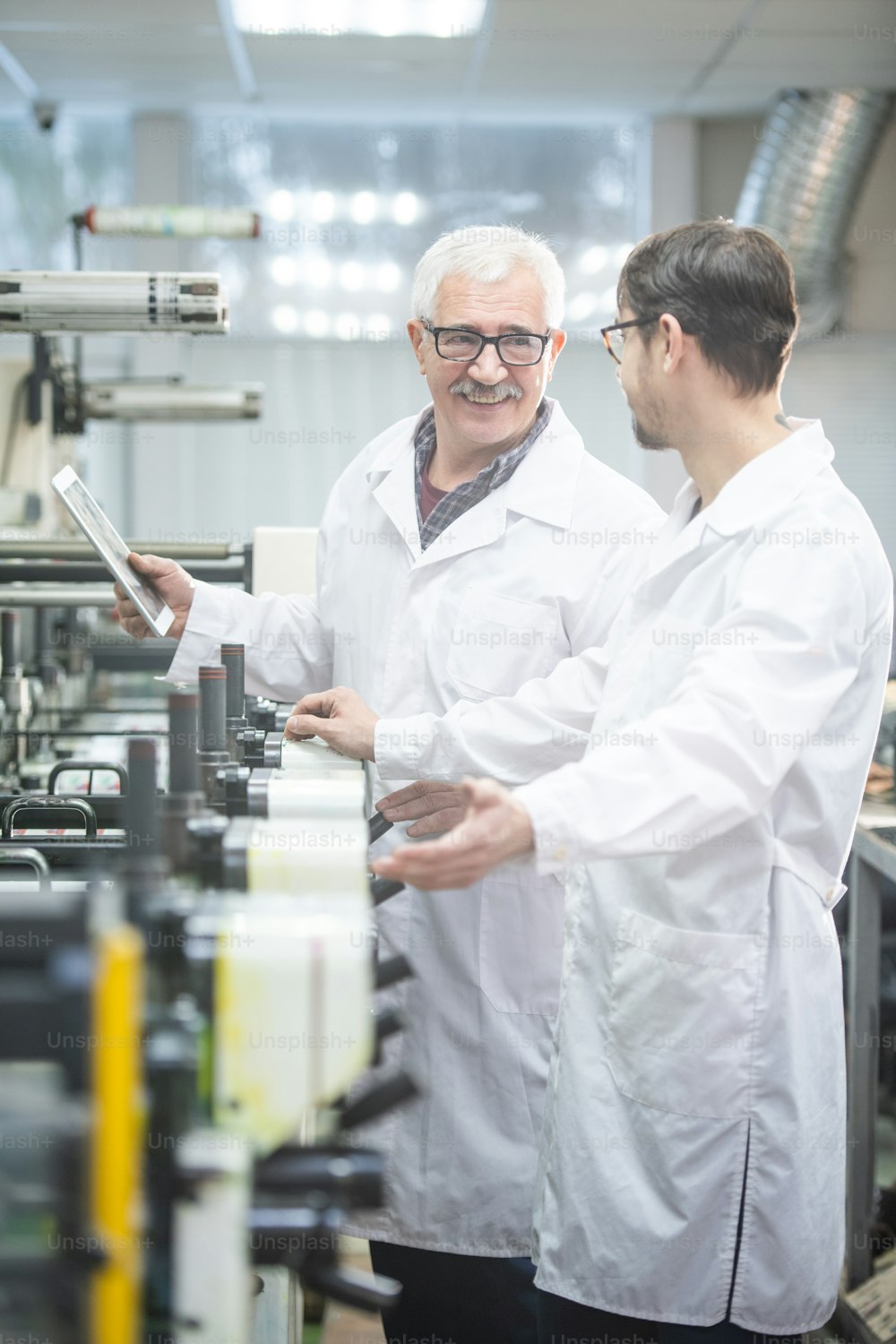 Smiling white-haired worker in lab coat using tablet while analyzing printing press productivity with colleague in factory shop