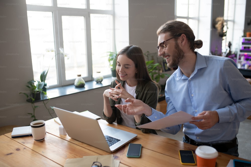 Business partners. Man with glasses and light blue shirt and girl with long dark hair working at a laptop in the office, smiling and joyful in good mood.