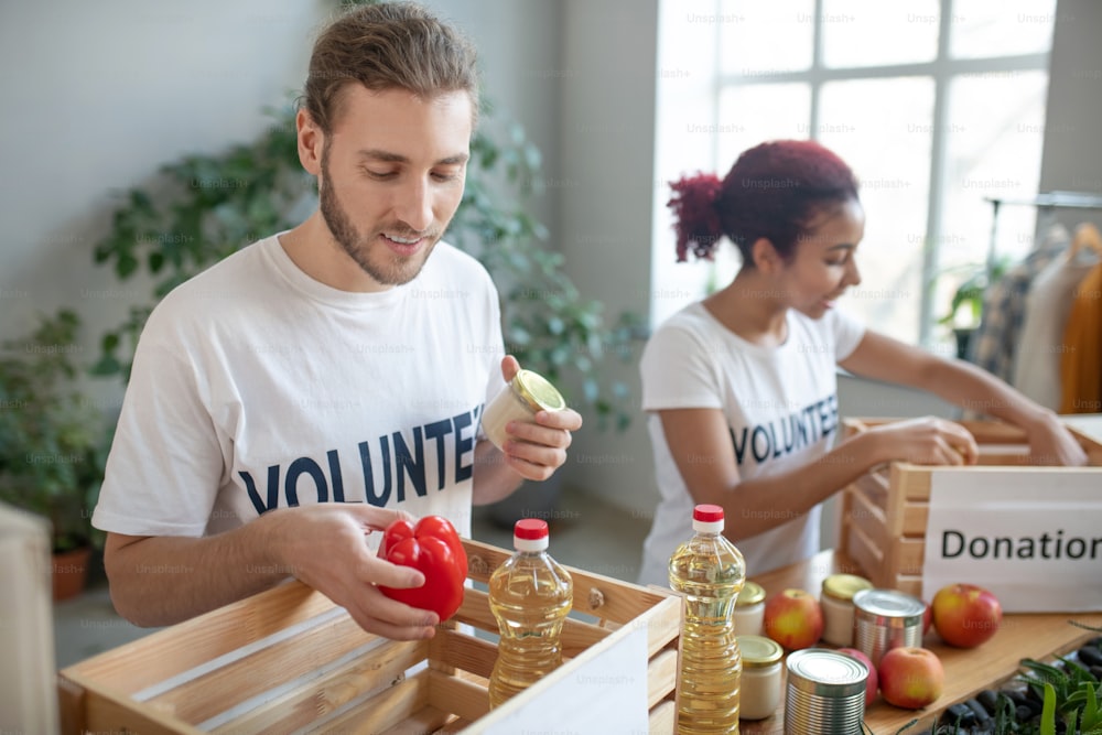 Good hearts. Young bearded man and girl with colored hair in white t-shirts standing together packing donation boxes, in good mood.