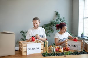 Packing process. Young girl with burgundy hair and a bearded man, standing at the table and packing up donation boxes with sets of products, cheerful smiling.