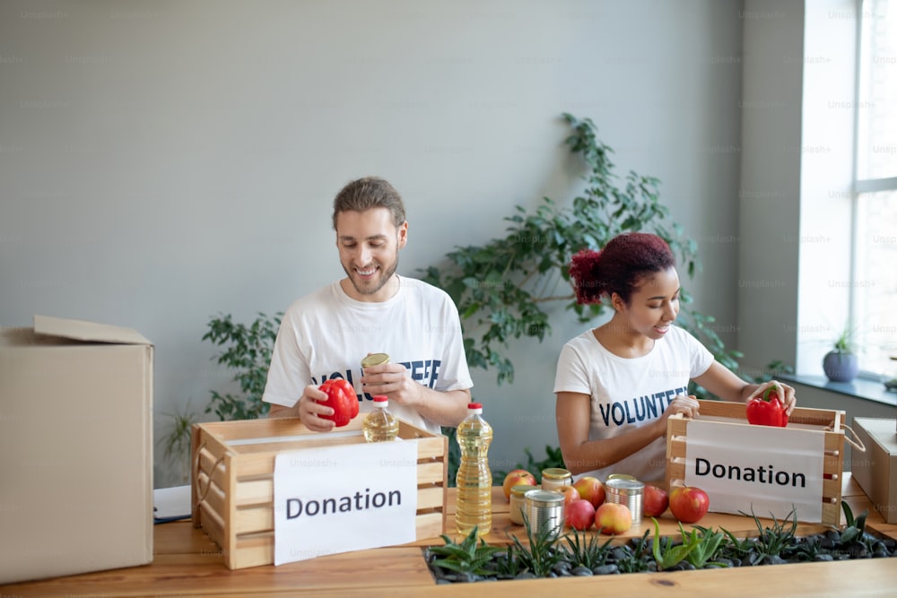 Packing process. Young girl with burgundy hair and a bearded man, standing at the table and packing up donation boxes with sets of products, cheerful smiling.