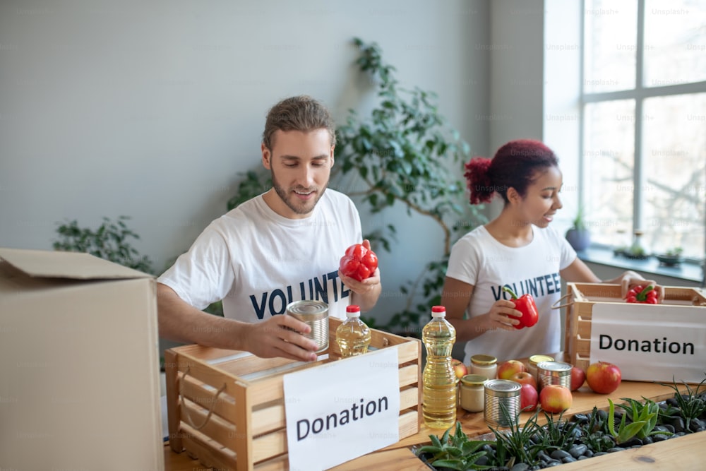 Teamwork. Young man with a can of canned food and pepper in his hands, a girl putting red peppers in a donation box, working harmoniously and joyfully.
