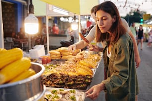 Toma horizontal de una joven pareja caucásica que pasa la noche en el mercado asiático de comida callejera eligiendo comida