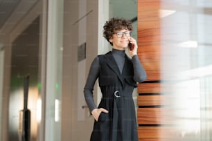 Portrait of smiling attractive young businesswoman in eyeglasses and black dress standing in corridor and talking to client on phone