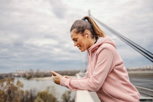 Side view of attractive smiling positive young sportswoman leaning on bridge railing, using smart phone for reading message and listening music. Urban life concept.