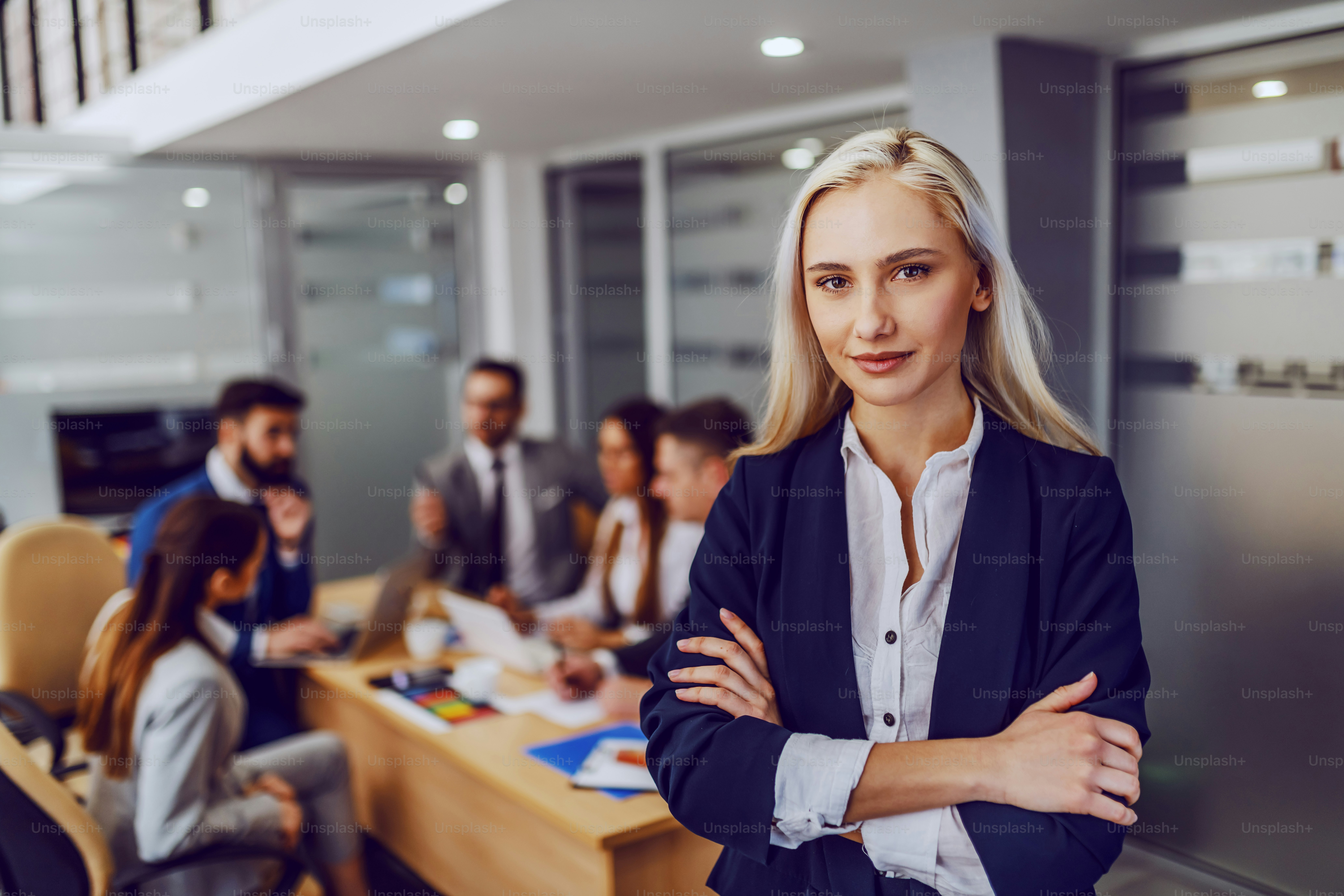 Junge lächelnde, erfolgreiche, wunderschöne blonde Geschäftsfrau in formeller Kleidung, die mit verschränkten Armen im Sitzungssaal steht und in die Kamera schaut. Im Hintergrund ihre Kollegen, die sich treffen.