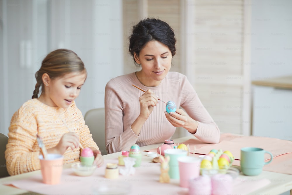 Warm-toned portrait of smiling young mother and daughter painting Easter eggs in cozy kitchen interior, copy space