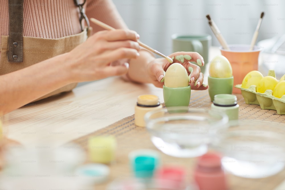 부엌이나 미술 스튜디오의 테이블에 앉아 부활절을 위해 파스텔 색상으로 달걀을 칠하는 알아볼 수 없는 젊은 여성의 클로즈업, 복사 공간