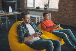 Muy relajante. Hombres felices y relajados sentados en las sillas de pufs mientras juegan videojuegos.