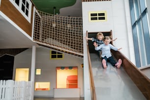 Two cute cheerful kids in casualwear moving down slide while having fun on play area in children center