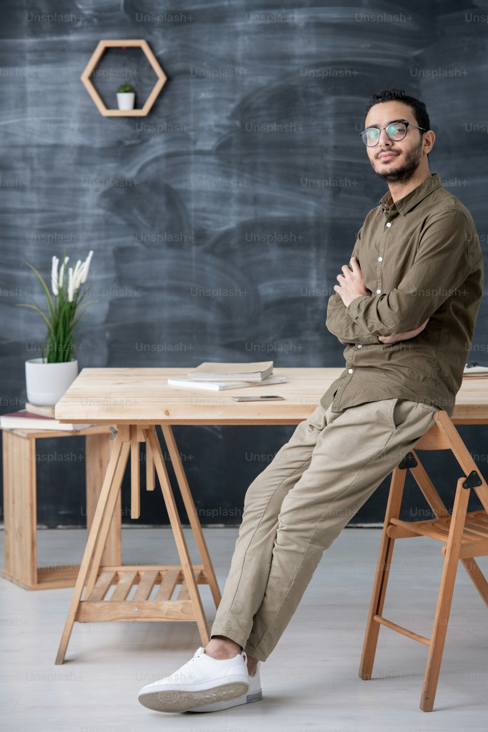 Young confident businessman or office worker crossing arms on chest while standing by table on background of blackboard