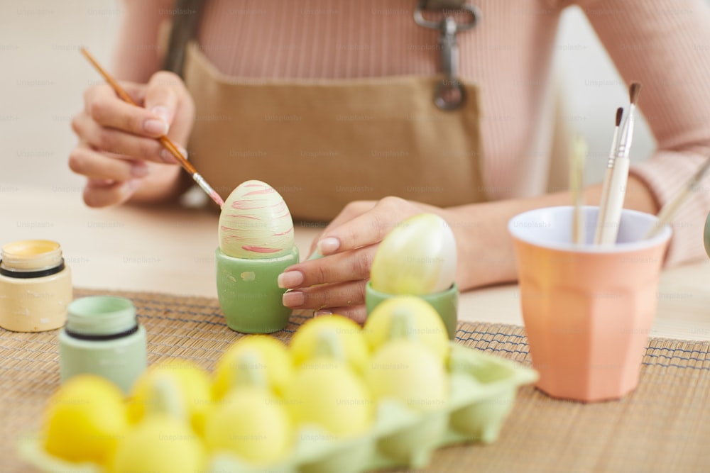 Close up de mulher jovem pintando ovos em cores pastel para a Páscoa enquanto está sentada à mesa na cozinha ou estúdio de arte, espaço de cópia