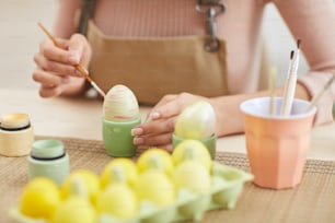 Close up of young woman painting eggs in pastel colors for Easter while sitting at table in kitchen or art studio, copy space
