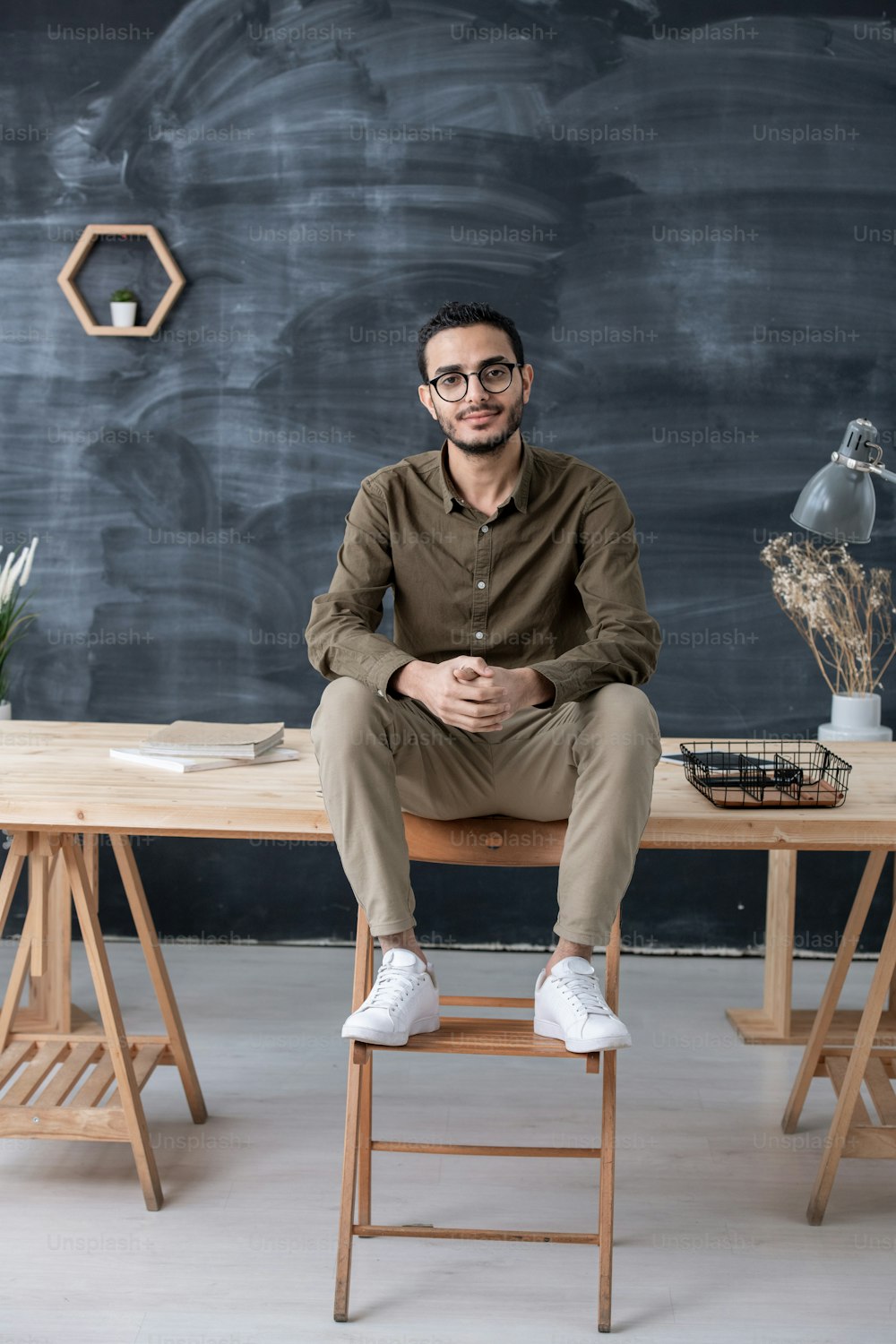 Jeune homme d’affaires confiant en tenue décontractée qui vous regarde assis sur table avec ses jambes sur une chaise en bois