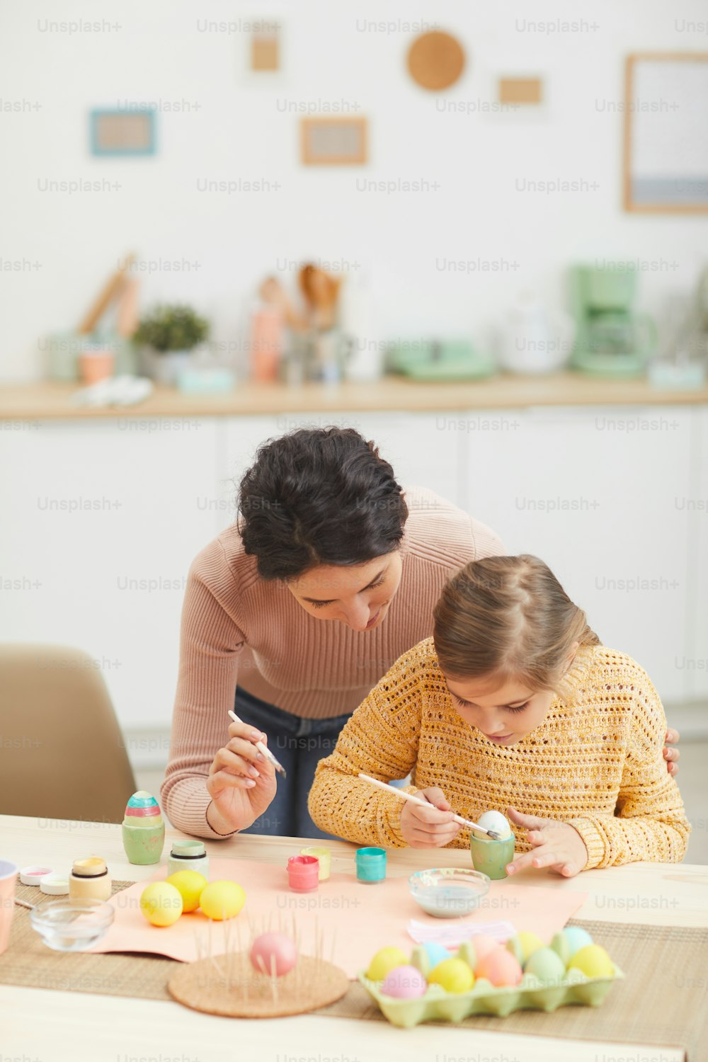 居心地の良いキッチンインテリアのテーブルに座ってイースターエッグを描く母と娘の垂直ポートレート、コピー用スペース