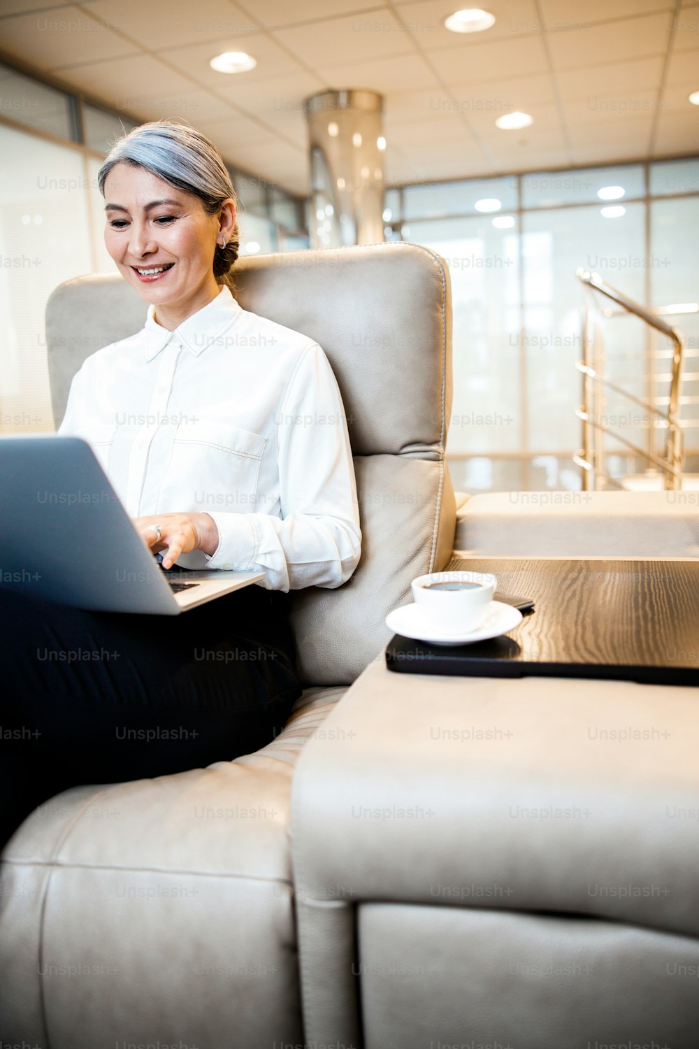 Enthusiastic entrepreneur sitting in a comfortable armchair with a cup of coffee by her side and using a modern laptop