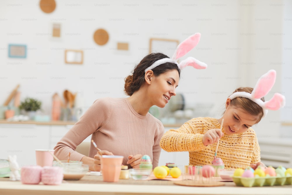 居心地の良いキッチンインテリアでイースターエッグを描くのを楽しむかわいい女の子を持つ若い母親のポートレート、両方ともウサギの耳、コピー用スペース