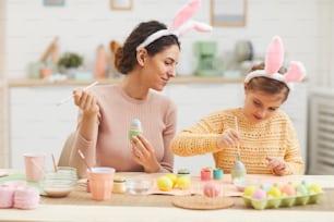Portrait d’une jeune mère avec une petite fille s’amusant tout en peignant des œufs de Pâques dans un intérieur de cuisine confortable, tous deux portant des oreilles de lapin, espace de copie