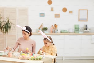 Ritratto di giovane madre e figlia che dipingono uova di Pasqua in interni accoglienti della cucina, entrambi indossano orecchie da coniglio, spazio copia