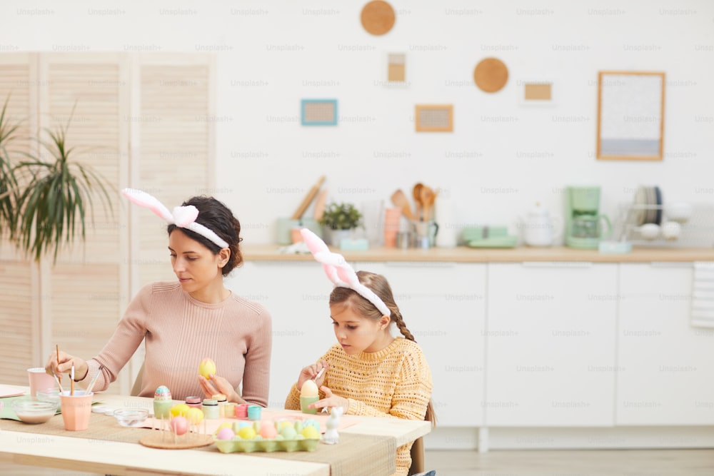 居心地の良いキッチンインテリアにイースターエッグを描く若い母と娘のポートレート、両方ともウサギの耳、コピー用スペースを着ている