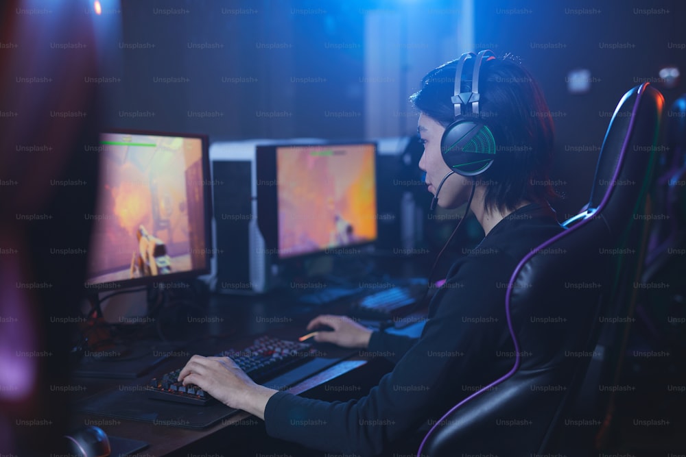 Ritratto laterale del giovane asiatico che gioca ai videogiochi e indossa le cuffie in interni informatici scuri, spazio di copia