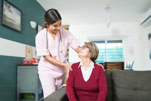Joven enfermera sonriente hablando con una mujer mayor sentada en el sofá de su casa