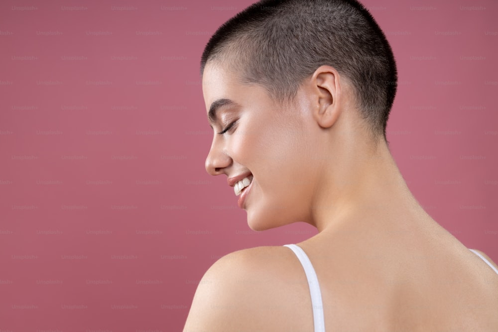 Attractive short haired young girl standing bare shouldered against the pink background and smiling