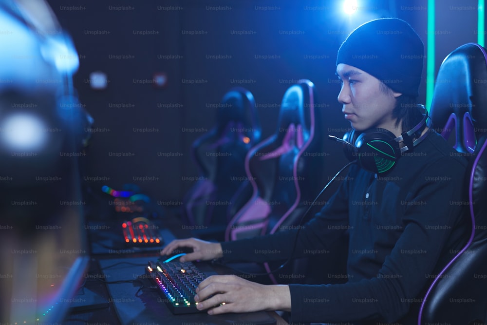 Seitenansichtsporträt eines jungen asiatischen Mannes, der Videospiele in dunklem cyberartigem Innenraum und Kopierraum spielt