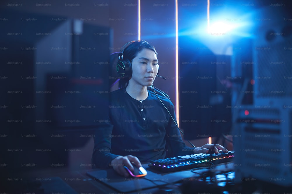 Retrato do jovem asiático usando o computador no interior escuro do cyber com o brilho da lente, espaço da cópia