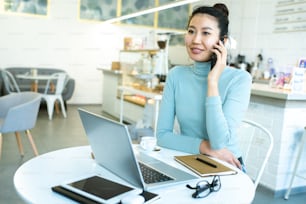 Hübsche junge lächelnde Geschäftsfrau oder Studentin, die sich mit dem Smartphone unterhält, während sie sich am Tisch im Café vor dem Laptop entspannt