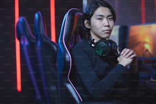 Retrato do jovem asiático olhando para a câmera enquanto está sentado na cadeira de jogos no interior escuro do esporte cibernético, espaço da cópia