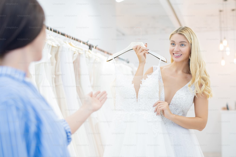 Excited beautiful bride with curly hair showing wedding dress to friend in shop