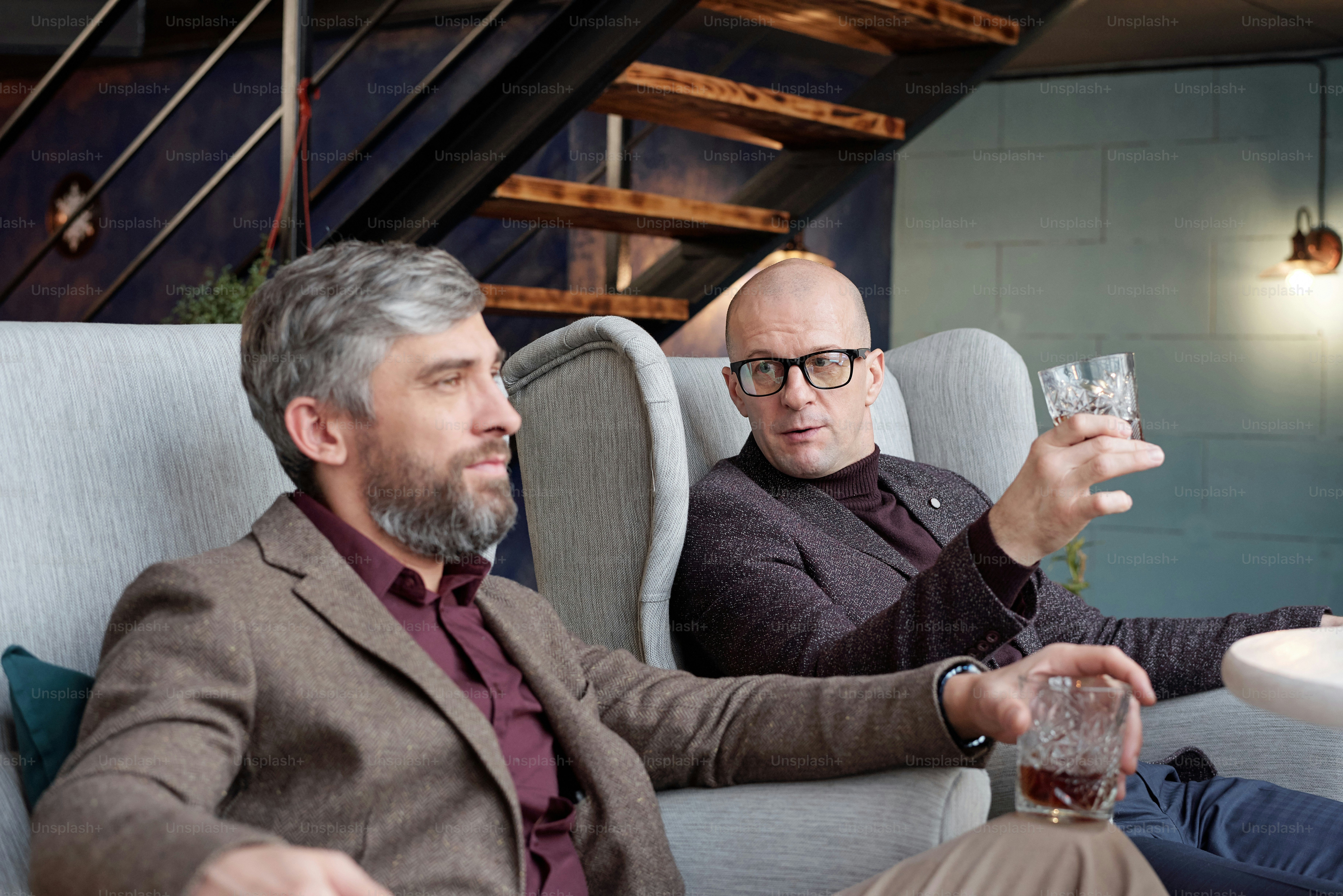 Bald businessman with whisky glass sitting in living room and discussing work with colleague