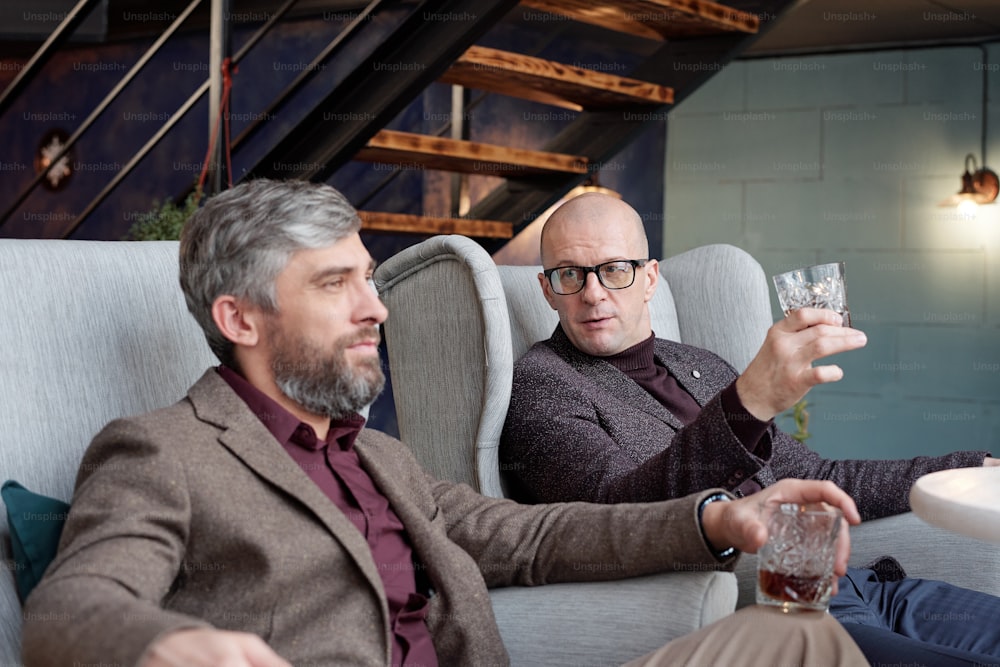 Bald businessman with whisky glass sitting in living room and discussing work with colleague