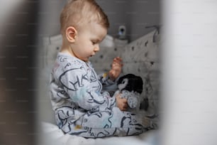 Retrato de un encantador bebé rubio sentado en su cuna y jugando con su juguete de pingüino favorito. Horario de la mañana.