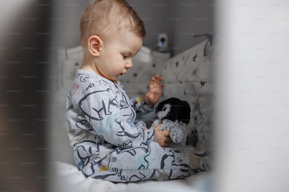 Portrait of charming blond little baby boy sitting in his crib and playing with his favorite penguin toy. Morning time.