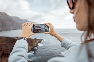 Viaggiatrice donna che fotografa sul telefono panorami mozzafiato sulla costa rocciosa dell'oceano mentre viaggia sull'isola di Tenerife, Spagna