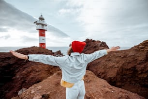 Femme élégante et insouciante vêtue d’un jean et d’un chapeau rouge profitant d’un voyage sur un rivage rocheux de l’océan près du phare, voyageant au nord-ouest de l’île de Tenerife, en Espagne