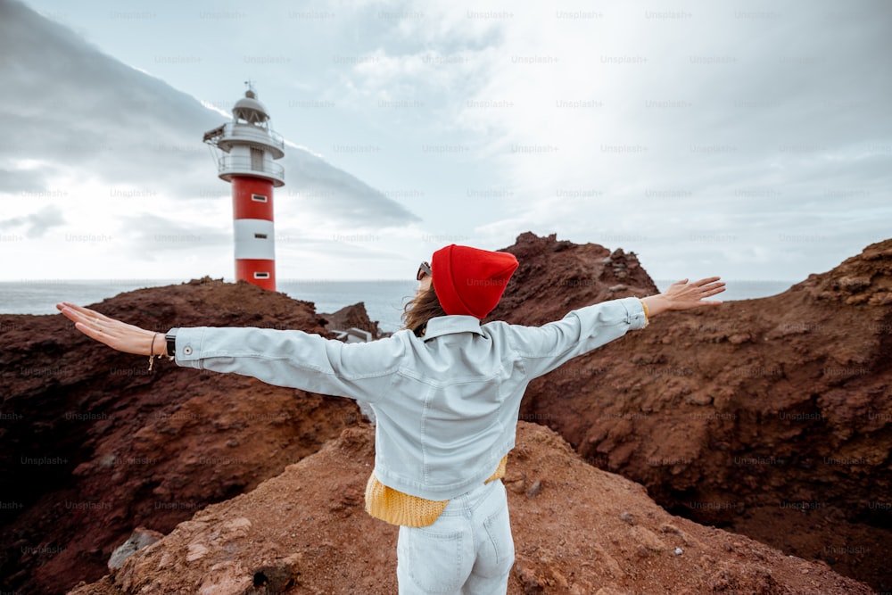 Mulher elegante despreocupada vestida com jeans e chapéu vermelho desfrutando de viagem em uma costa rochosa do oceano perto do farol, viajando no noroeste da ilha de Tenerife, Espanha